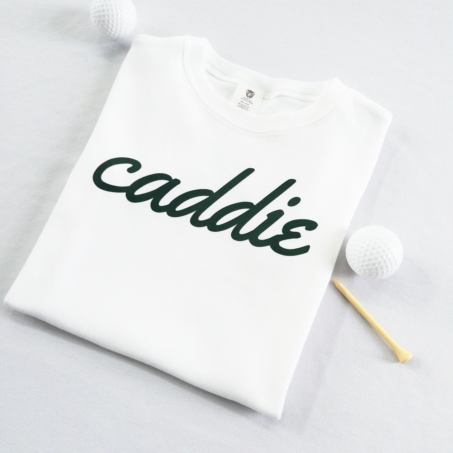 White kids golf t-shirt with dark green cursive text reading 'caddie,' displayed flat with golf balls and a wooden tee, perfect for young golf enthusiasts and future caddies.