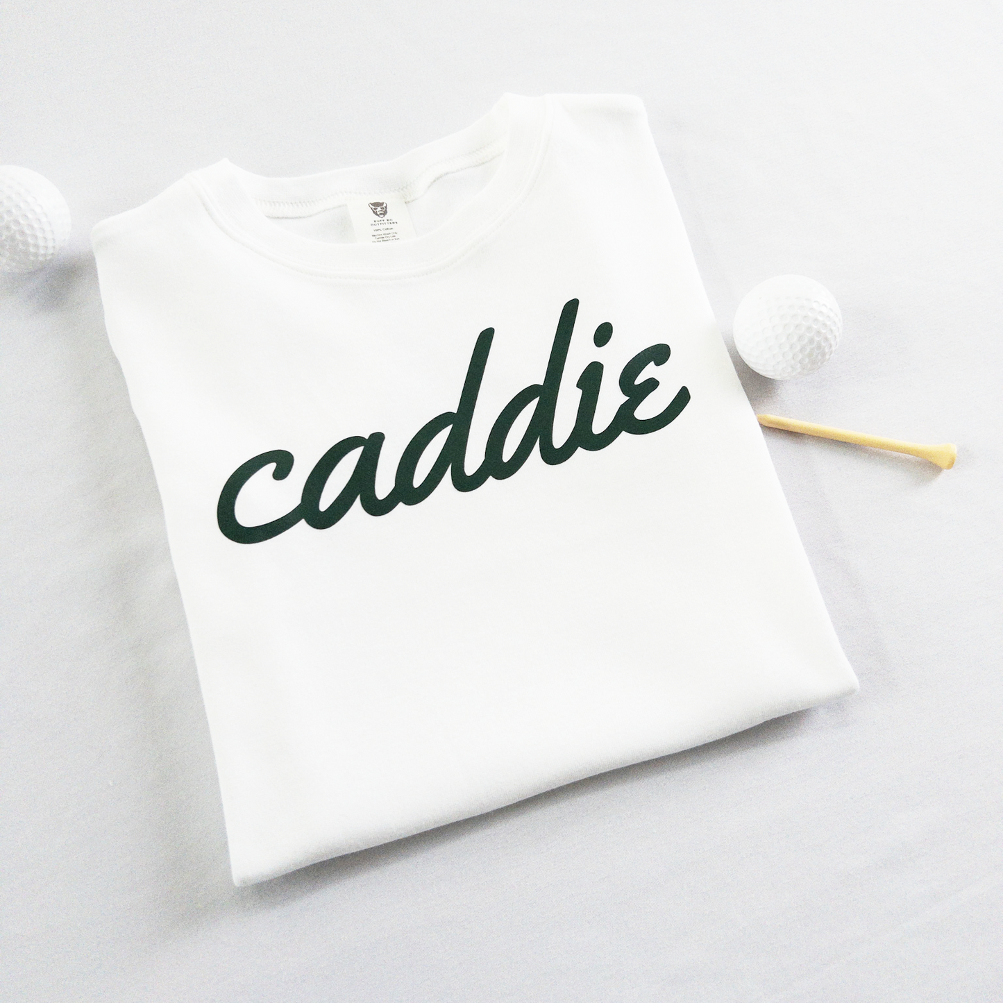 White kids golf t-shirt with dark green cursive text reading 'caddie,' displayed flat with golf balls and a wooden tee, perfect for young golf enthusiasts and future caddies.