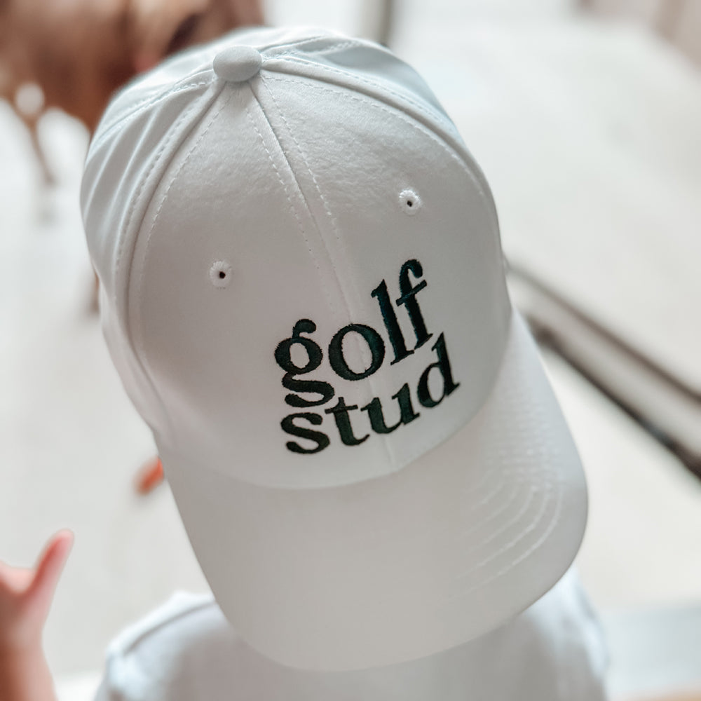Child wearing a white baseball cap with bold green embroidered 'golf stud' text, casual and playful golf hat perfect for kids and youth golfers, photographed indoors with soft natural light.