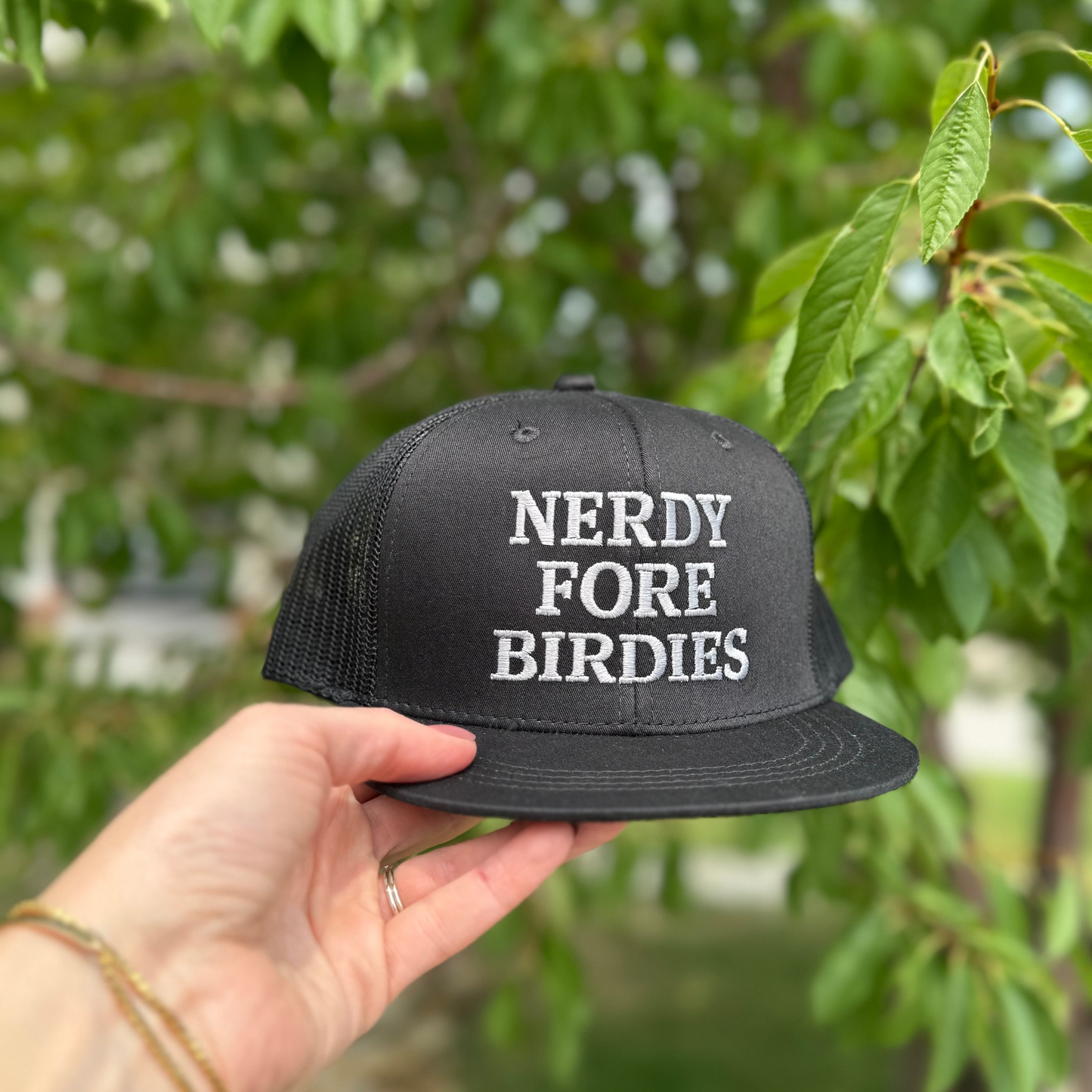 Black cap with 'NERDY FORE BIRDIES' text held by a hand against a blurred green background