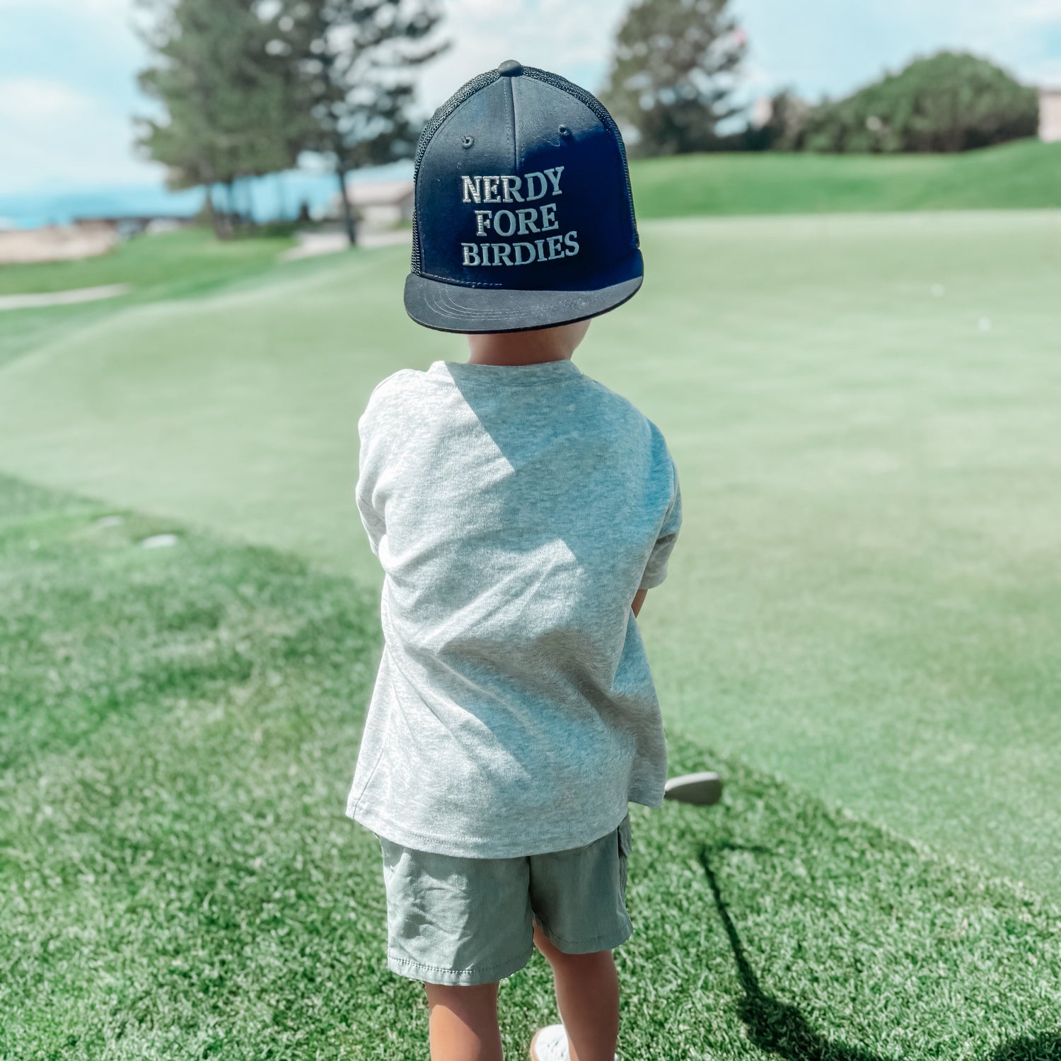 Child on a golf course wearing a cap with 'Nerdy Fore Birdies' text