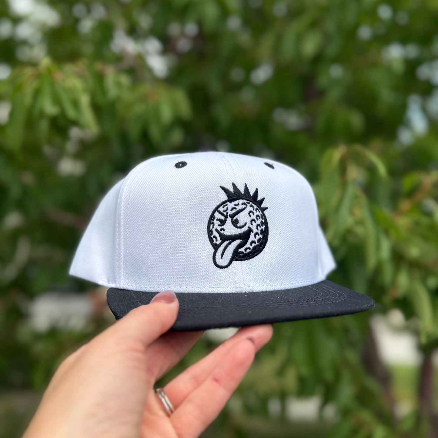 White cap with black logo held by a hand against a blurred green background