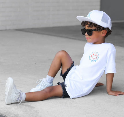 Child sitting on a concrete surface wearing a white t-shirt, black shorts, white sneakers, and sunglasses.