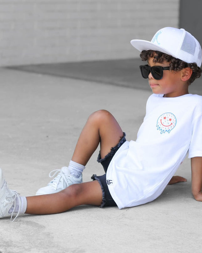Child sitting on a concrete surface wearing a white t-shirt, black shorts, white sneakers, and sunglasses.