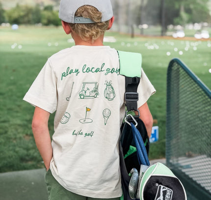 Child wearing a 'play local golf' t-shirt with a golf bag and club on a golf course.