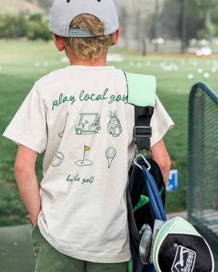 Child wearing a 'play local golf' t-shirt with a golf bag and club on a golf course.