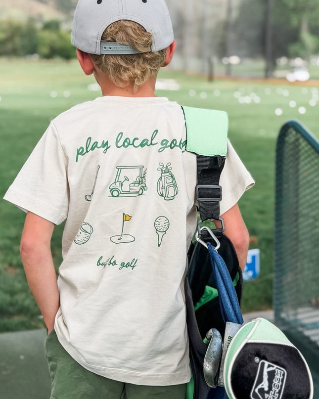Child wearing a 'play local golf' t-shirt with a golf bag and club on a golf course.