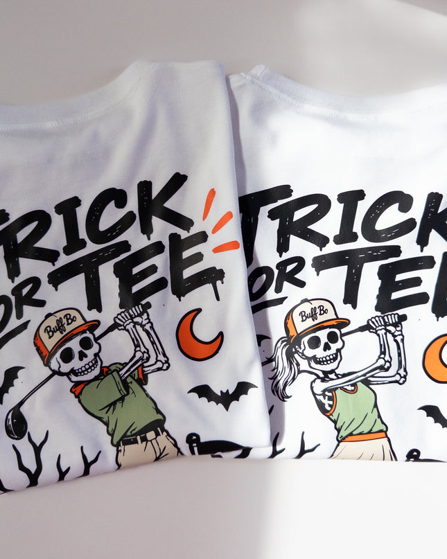 Two white short-sleeve T-shirts laid flat on a light gray surface, each printed with a Halloween-themed graphic that reads “TRICK OR TEE” in large black letters, with a cartoon skeleton, orange moon, bats, and black tree branches.
