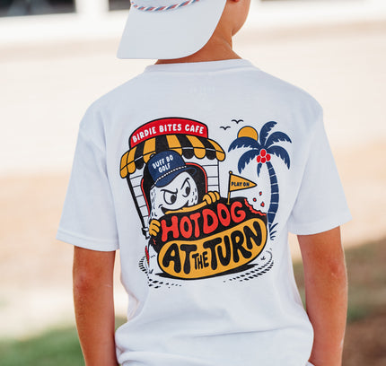 A child shown from the back wearing a white graphic t-shirt with a colorful cartoon hot dog vendor design that reads 'Hotdog at the Turn' and 'Dirt Bitz Cafe'. The child is also wearing a white baseball cap with an American flag pattern and camouflage shorts.