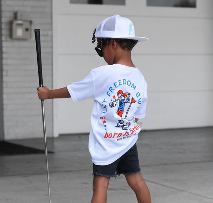 Child holding a golf club and wearing a 'Freedom' t-shirt in front of a garage.