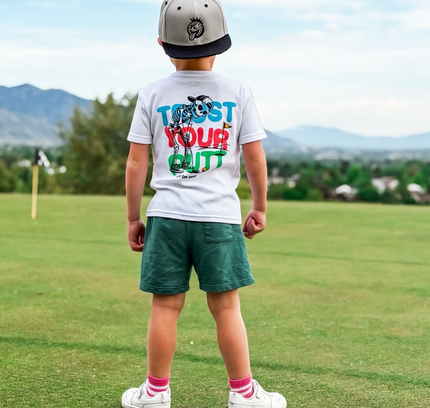 Child standing on a golf course wearing a white t-shirt with colorful text and graphics.