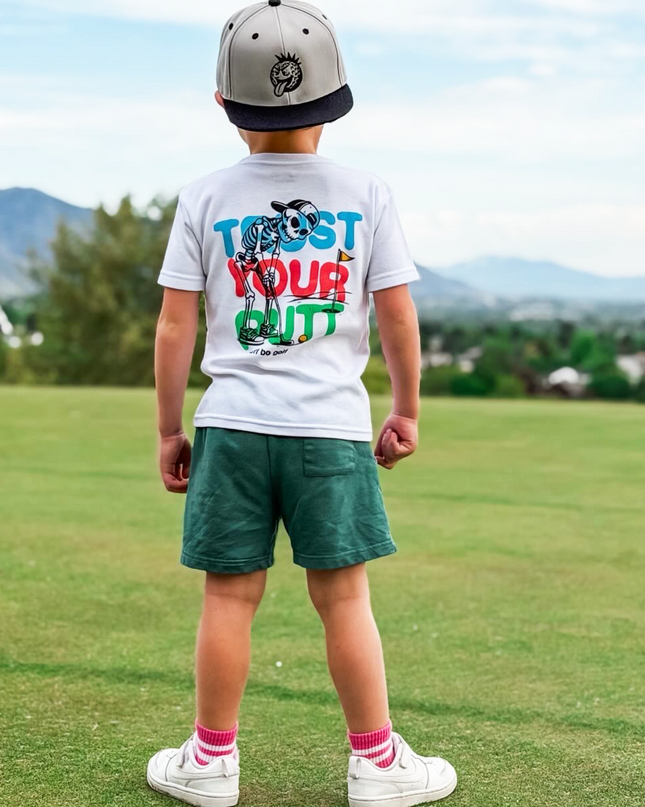 Child standing on a golf course wearing a white t-shirt with colorful text and graphics.