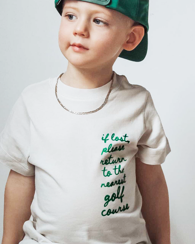 Child wearing a white t-shirt with text and a green cap on a plain background