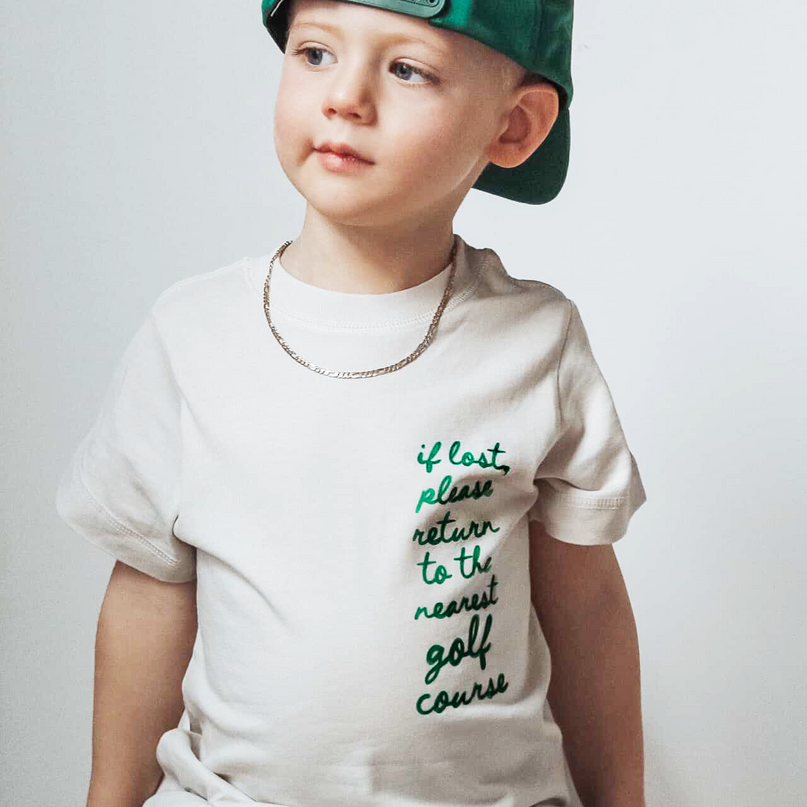 Child wearing a white t-shirt with text and a green cap on a plain background