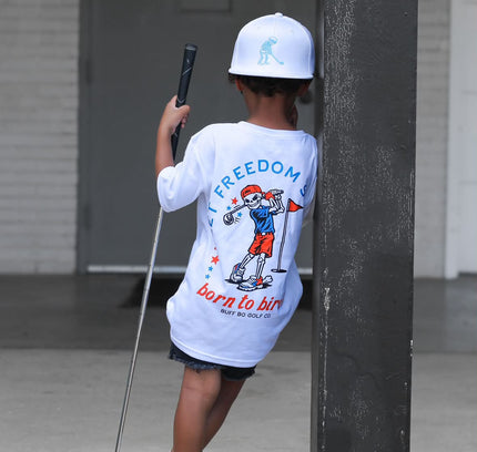 Child wearing a 'Born to Ski' t-shirt holding a ski pole, standing next to a building.