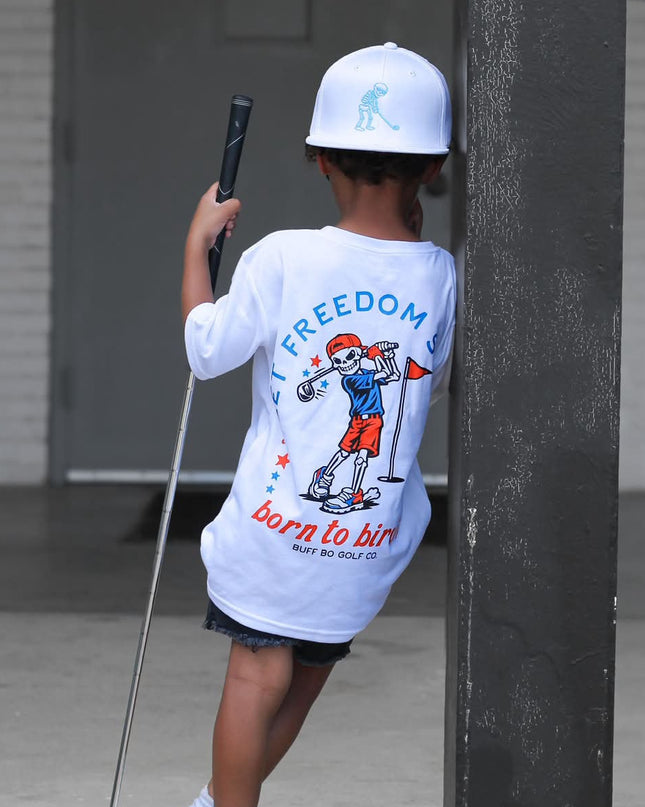 Child wearing a 'Born to Ski' t-shirt holding a ski pole, standing next to a building.