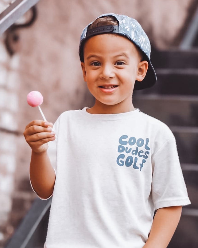 A young boy wearing a white graphic t-shirt with 'Cool Dudes Golf' printed in blue, khaki cargo pants, and a blue patterned baseball cap backwards, holding a pink lollipop while standing on outdoor stairs.