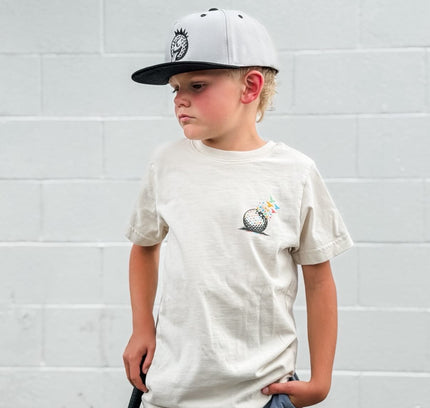Young boy wearing a cap and t-shirt, holding a golf club against a white wall.