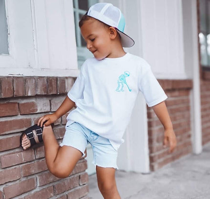 Child wearing a white t-shirt with a graphic design, light blue shorts, and a cap, standing against a brick wall.