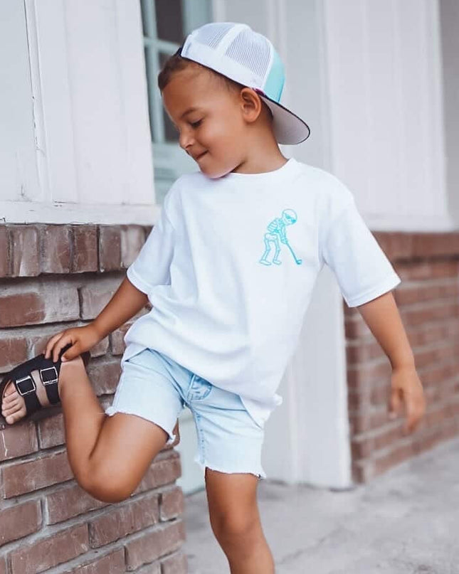 Child wearing a white t-shirt with a graphic design, light blue shorts, and a cap, standing against a brick wall.
