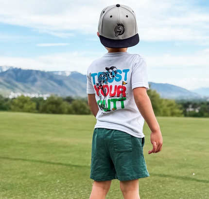 Child wearing a white t-shirt with colorful text and graphics, green shorts, and a cap, standing on grass with mountains in the background.