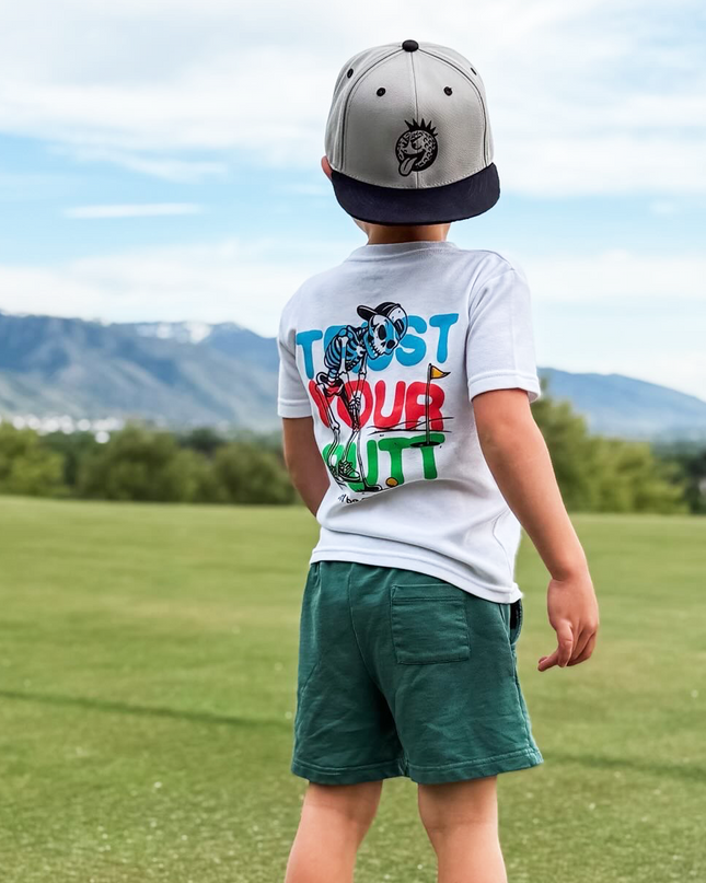Child wearing a white t-shirt with colorful text and graphics, green shorts, and a cap, standing on grass with mountains in the background.