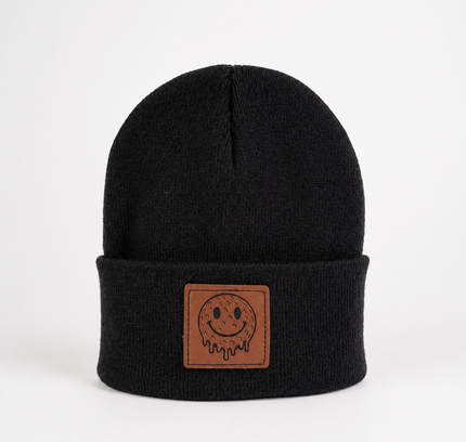 A black knit beanie hat with a folded cuff featuring a brown rectangular patch with a simple smiley face design, photographed against a plain white background.