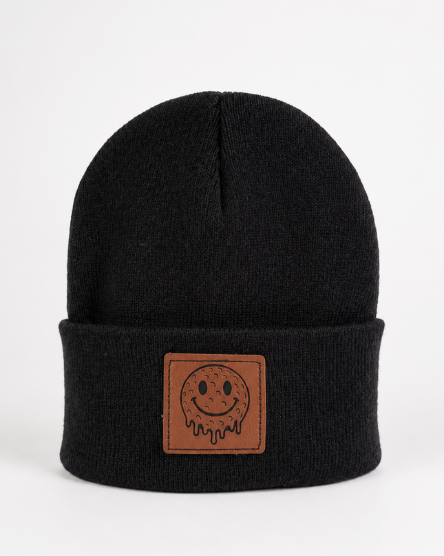 A black knit beanie hat with a folded cuff featuring a brown rectangular patch with a simple smiley face design, photographed against a plain white background.