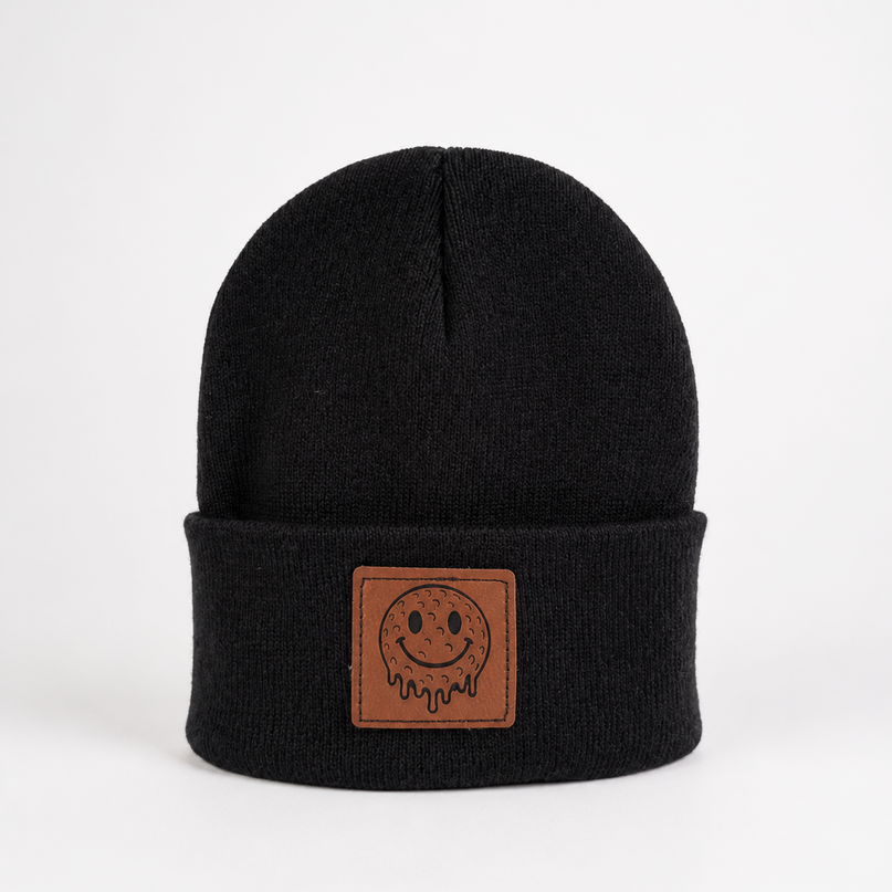 A black knit beanie hat with a folded cuff featuring a brown rectangular patch with a simple smiley face design, photographed against a plain white background.