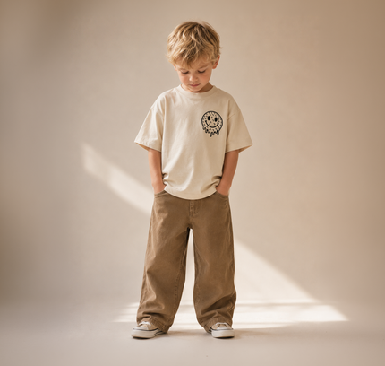 Child wearing a beige t-shirt and brown pants standing against a plain background