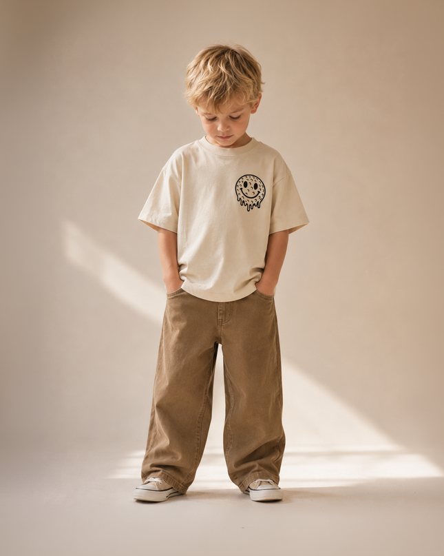 Child wearing a beige t-shirt and brown pants standing against a plain background