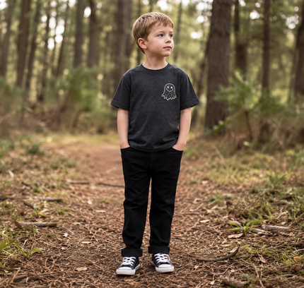 A young boy stands on a forest path wearing a dark charcoal gray t-shirt with a small white ghost graphic on the left chest, paired with black pants and black-and-white sneakers.