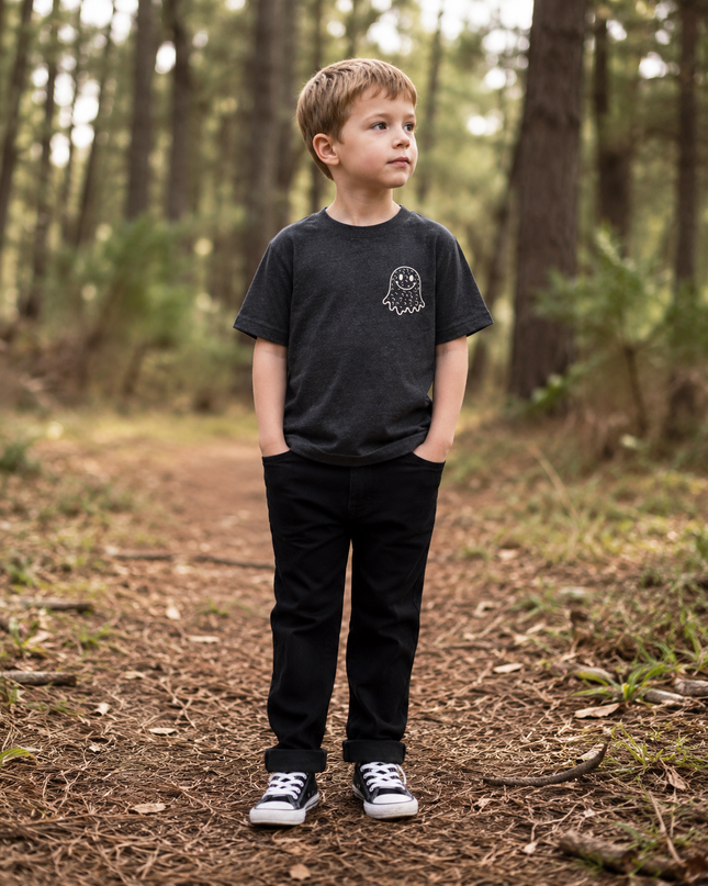 A young boy stands on a forest path wearing a dark charcoal gray t-shirt with a small white ghost graphic on the left chest, paired with black pants and black-and-white sneakers.