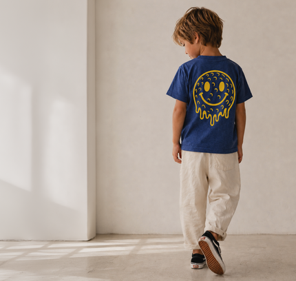 A child wearing a royal blue t-shirt with a yellow smiley face graphic on the back, paired with beige pants and black sneakers, walking away in a minimalist room with warm natural light.