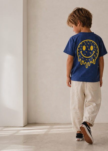 A child wearing a royal blue t-shirt with a yellow smiley face graphic on the back, paired with beige pants and black sneakers, walking away in a minimalist room with warm natural light.