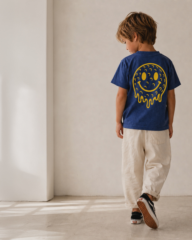 A child wearing a royal blue t-shirt with a yellow smiley face graphic on the back, paired with beige pants and black sneakers, walking away in a minimalist room with warm natural light.