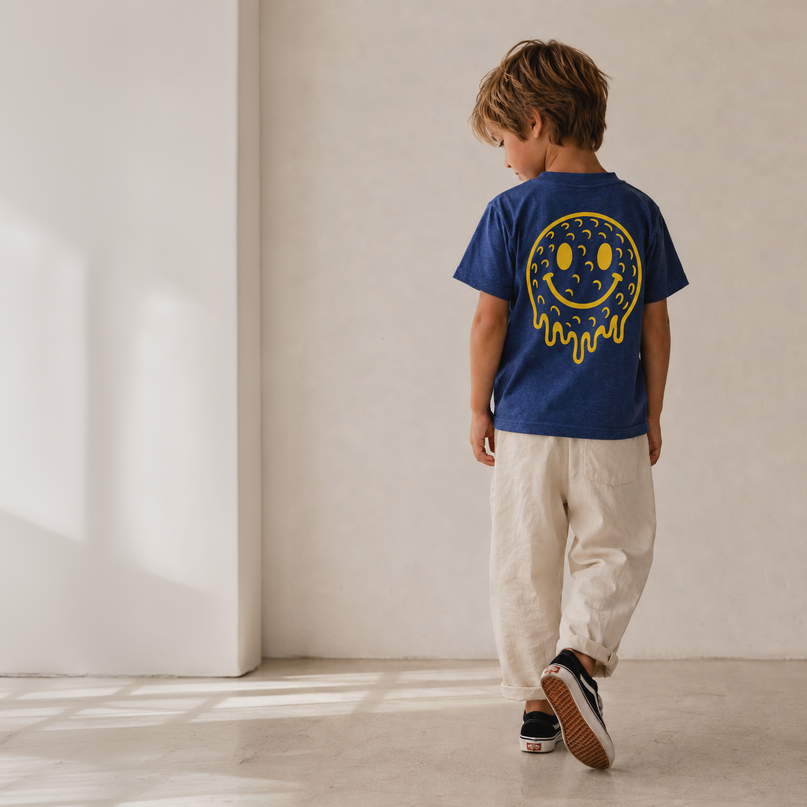 A child wearing a royal blue t-shirt with a yellow smiley face graphic on the back, paired with beige pants and black sneakers, walking away in a minimalist room with warm natural light.