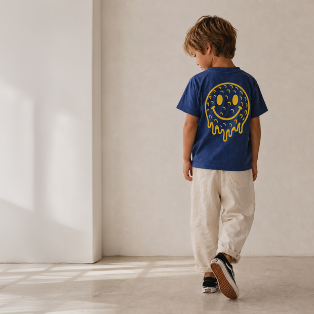 A child wearing a royal blue t-shirt with a yellow smiley face graphic on the back, paired with beige pants and black sneakers, walking away in a minimalist room with warm natural light.