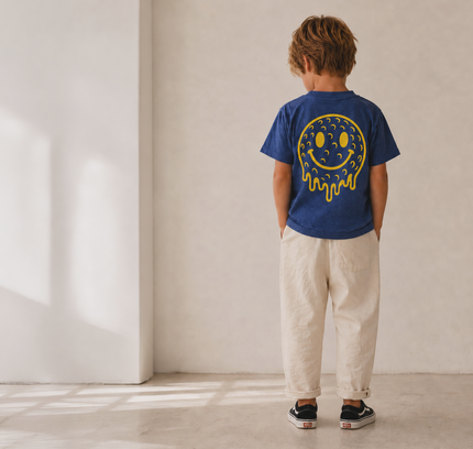 A child standing with their back to the camera in a minimalist room, wearing a royal blue t-shirt with a yellow dripping smiley face graphic and cream-colored pants.