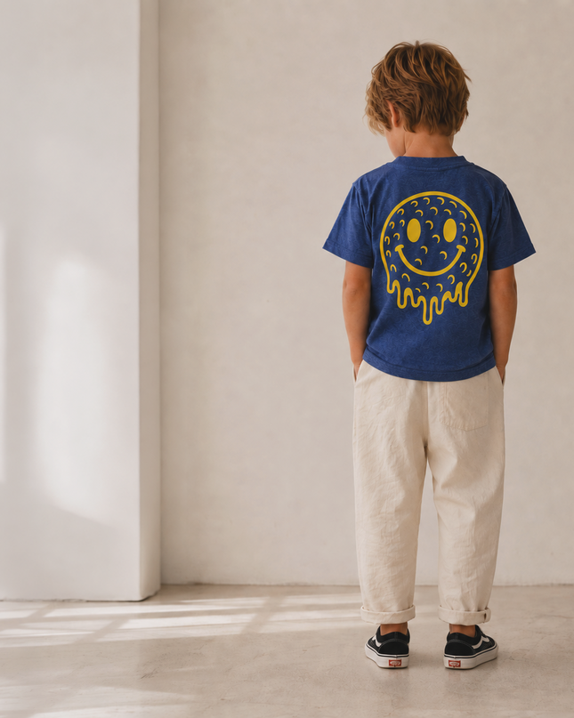 A child standing with their back to the camera in a minimalist room, wearing a royal blue t-shirt with a yellow dripping smiley face graphic and cream-colored pants.