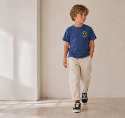 A young boy walking in a minimalist room with beige walls and concrete floors, wearing a blue crewneck t-shirt with a yellow smiley face graphic on the chest, beige trousers, and black low-top sneakers.