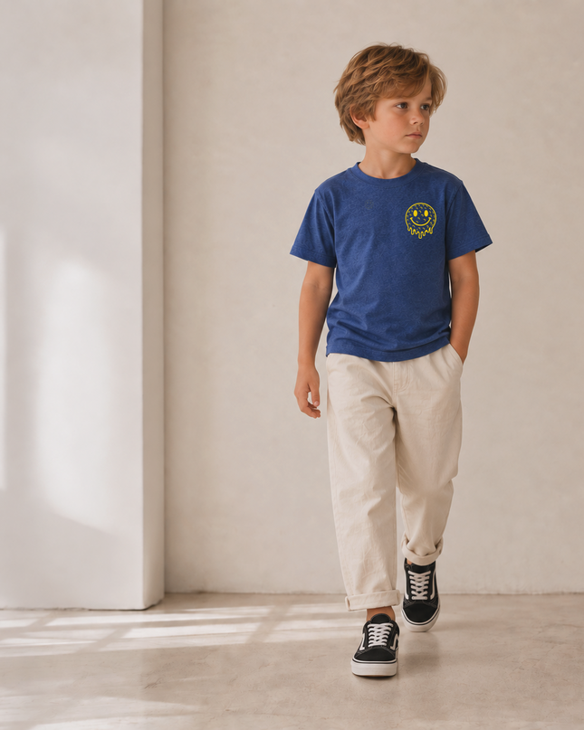 A young boy walking in a minimalist room with beige walls and concrete floors, wearing a blue crewneck t-shirt with a yellow smiley face graphic on the chest, beige trousers, and black low-top sneakers.