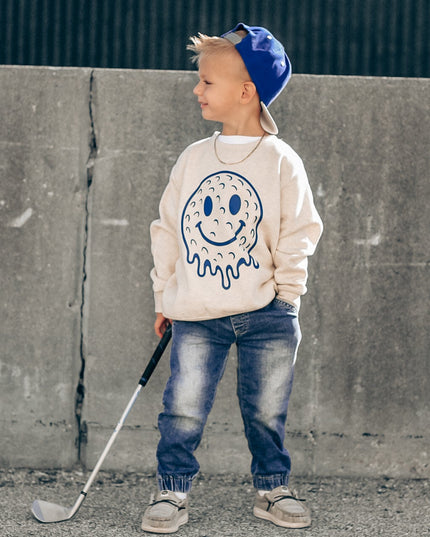 Child wearing a sweatshirt with a smiley face design, holding a golf club, standing against a concrete wall.