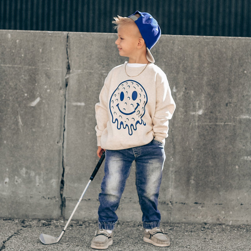 Child wearing a sweatshirt with a smiley face design, holding a golf club, standing against a concrete wall.