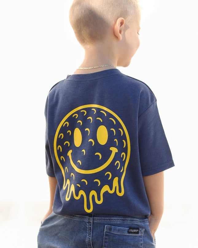 Child wearing a blue t-shirt with a yellow smiley face design on the back, sitting on a white surface.