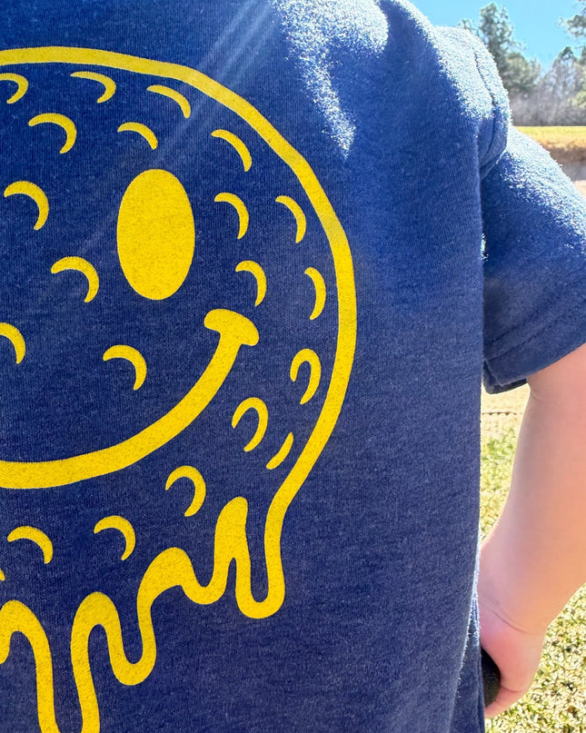 Person wearing a blue t-shirt with a yellow smiley face design outdoors.