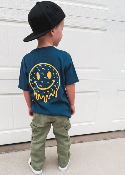 Child wearing a blue t-shirt with a smiley face design, standing in front of a white garage door.