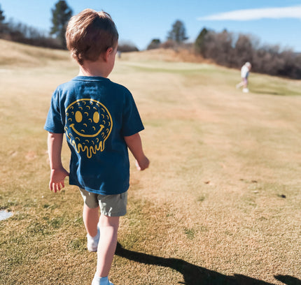 Child wearing a blue shirt with a smiley face design walking on a grassy field.