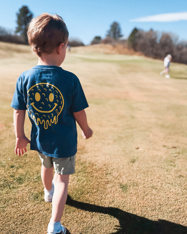 Child wearing a blue shirt with a smiley face design walking on a grassy field.