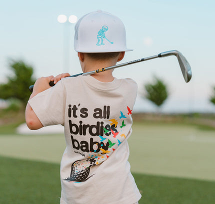 Child holding a golf club on a golf course wearing a t-shirt with text and graphics.